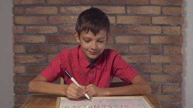 孩子坐着桌子上持有白板纸上的内容刻字签名背景红色的砖墙